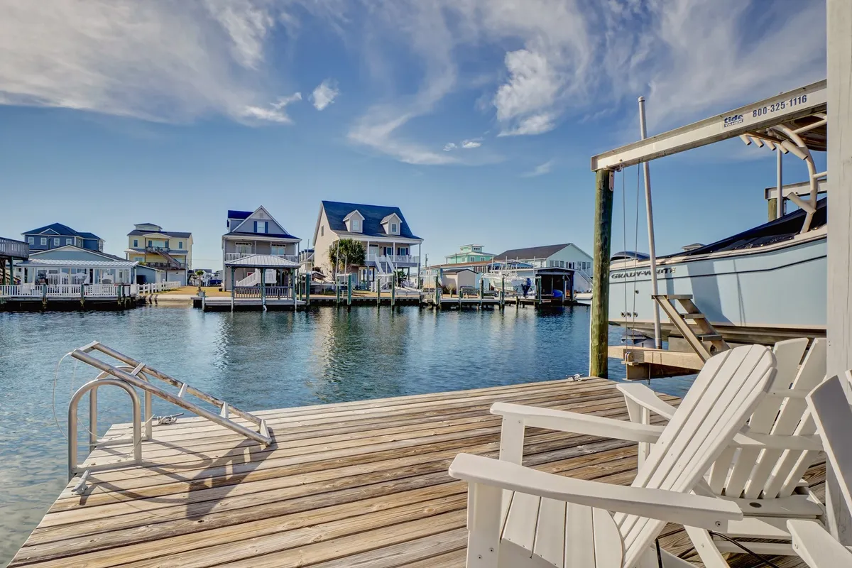 A peaceful wooden dock extending over the water, with adirondack chairs for relaxing, waterfront houses in the background, and a boat moored nearby. The tranquil mood invites fishing, sunbathing, and socializing by the canal.