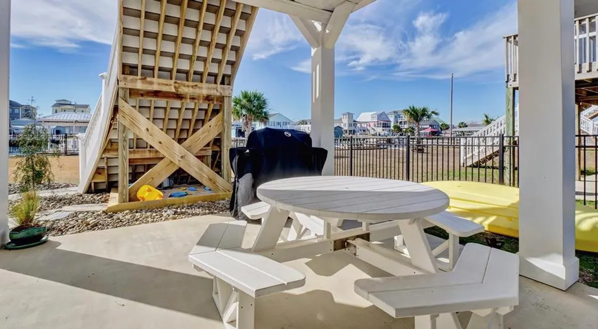 A playful and active backyard setting with a white picnic table, a wooden slide structure, and colorful kayaks ready for action. Palm trees and bright sunlight reinforce the relaxed, outdoor lifestyle.