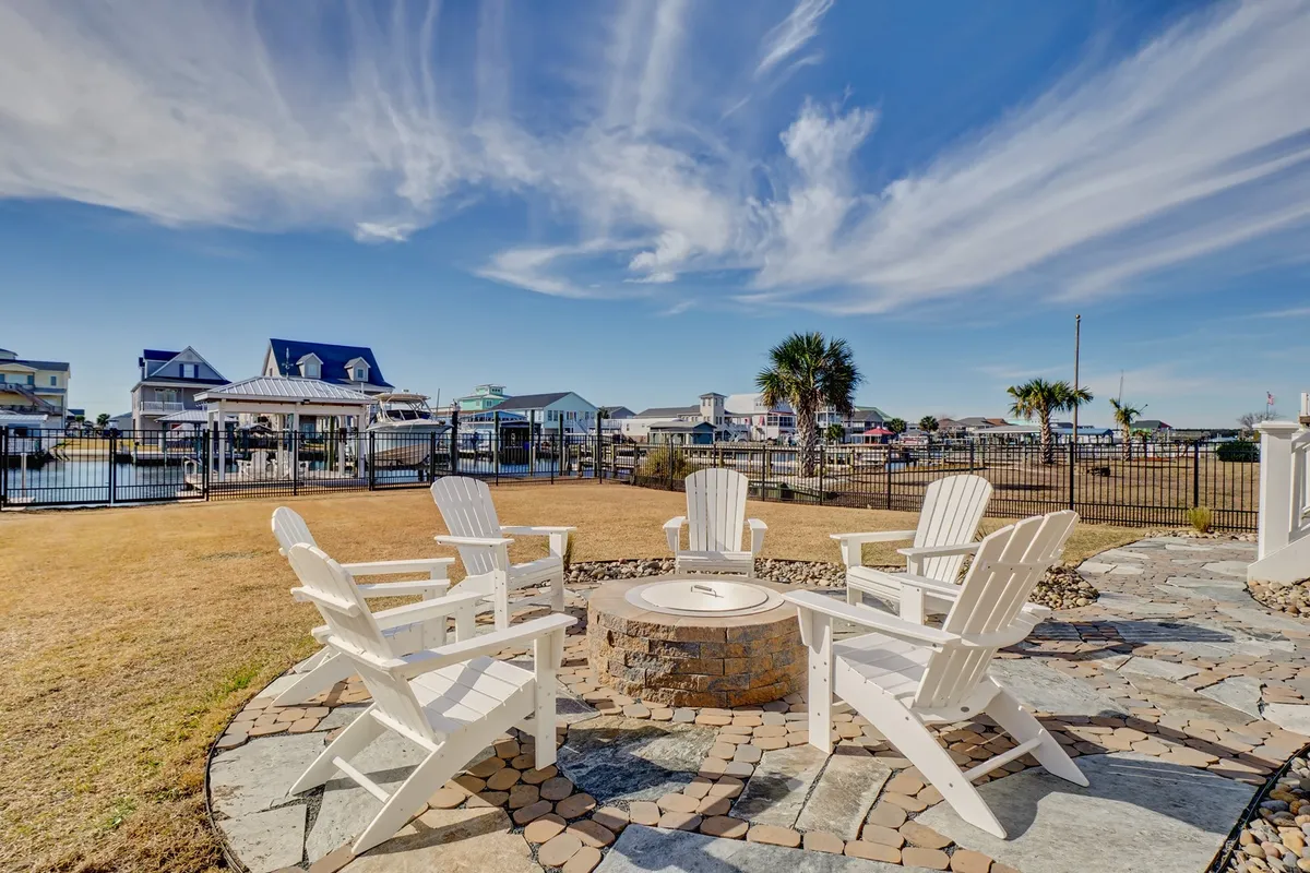An exterior seating area with Adirondack chairs set around a fire pit, all overlooking a serene water view and open skies. The mood is communal and relaxed, inviting gatherings under the stars or peaceful moments by the water.