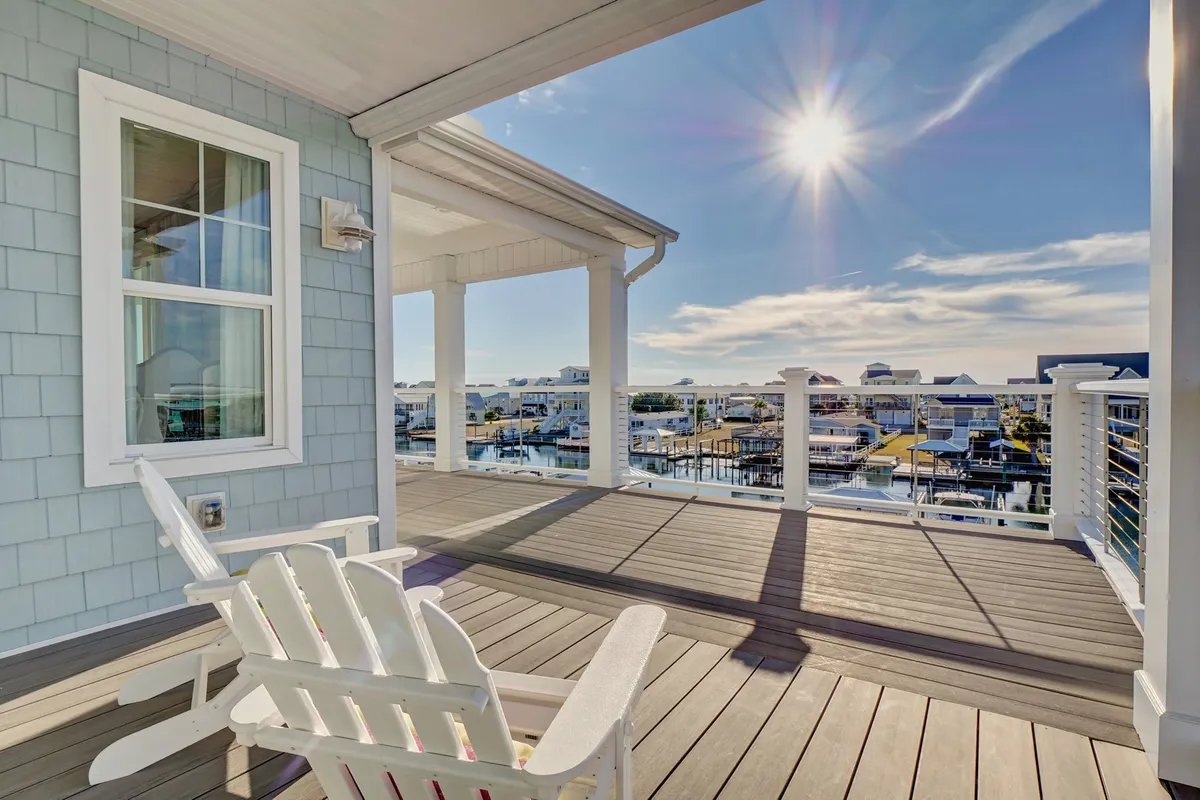An inviting outdoor deck features white Adirondack chairs arranged to face a waterfront view, with sunlight shimmering on the water. The scene exudes tranquility and sophistication, with the deck offering a luxurious space to relax, entertain, or enjoy sunrise and sunset vistas over the sound.