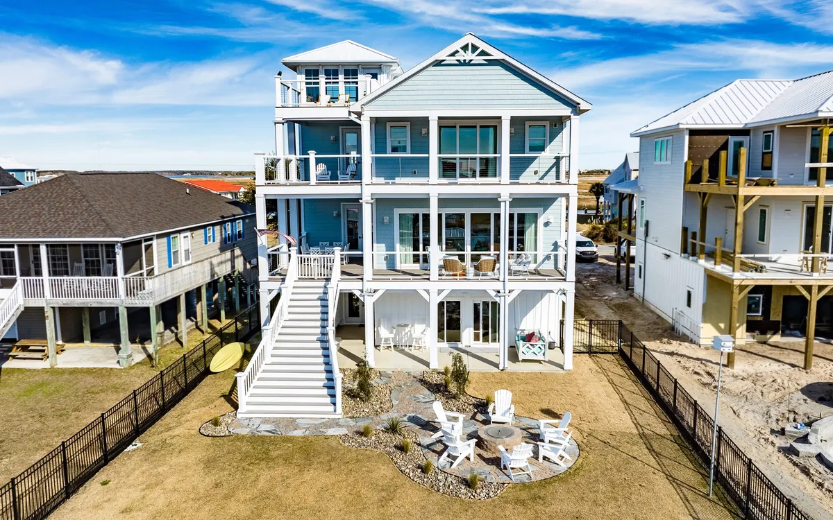 A multi-story beach house with an inviting outdoor seating area centered around a fire pit, surrounded by a landscaped yard and scenic open space. The home features expansive balconies and decks, creating plenty of areas for groups to gather and enjoy the outdoors together.