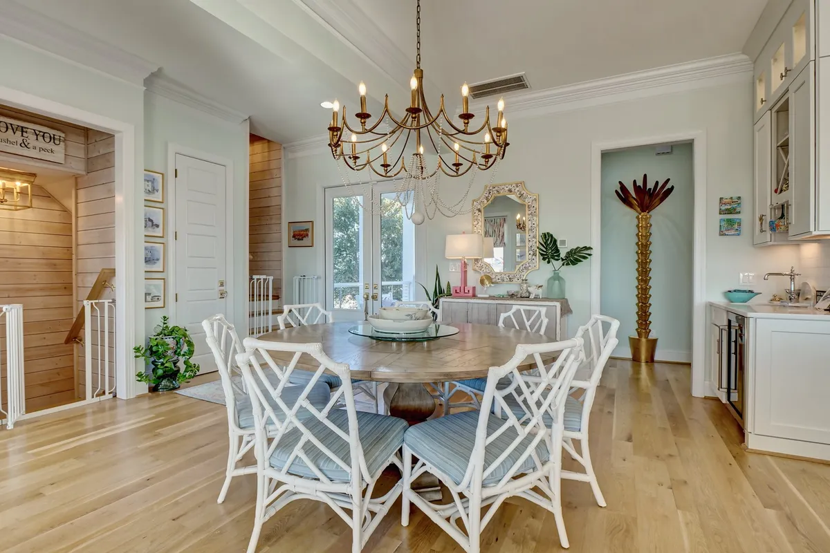 A warm, open dining area is shown with a central table beneath an elegant chandelier, surrounded by greenery, mirrors, and a view into the adjoining kitchen space. The setting is inviting and spacious, suitable for both intimate family meals and larger celebrations.