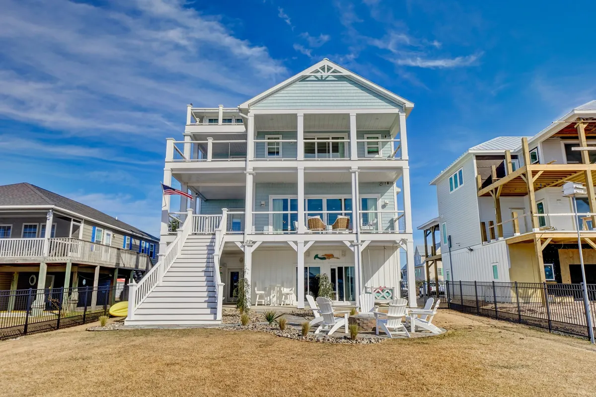 The three-story structure features wide decks and an expansive outdoor seating area, surrounded by a well-maintained lawn. Family and friends are pictured relaxing, dining, and celebrating together under the evening sky, with the ocean just beyond. The scene radiates warmth, space, and an effortless blend of indoor-outdoor living.