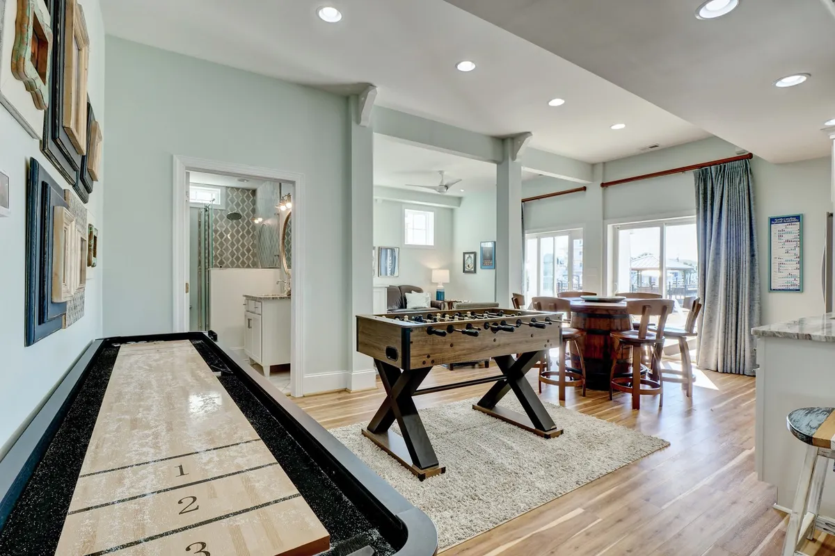 This image showcases a lively game room featuring a shuffleboard table and a foosball table, along with a barrel-style dining table and large windows offering plenty of natural light and views. The setup is ideal for friendly competitions and socializing, echoing the playful and inclusive spirit described in the section.