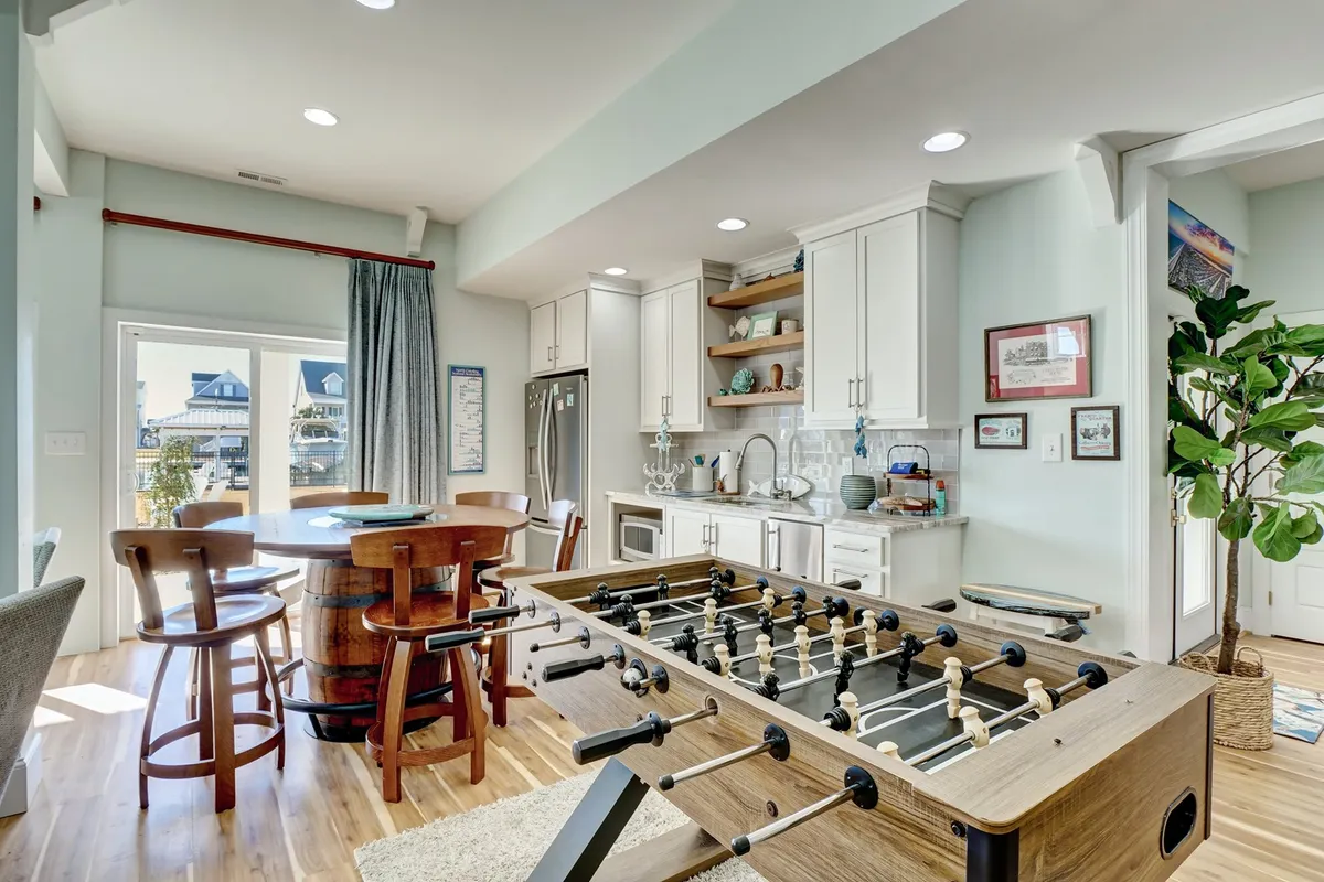 An inviting interior scene featuring a foosball table, a dining area, and kitchen cabinets, with plenty of natural light streaming in. The open, multi-purpose space is perfect for games, meals, and laughter-filled family moments.